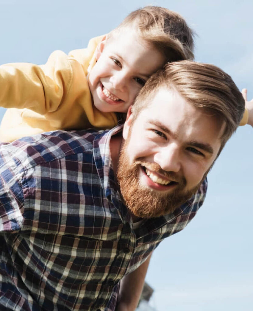 Ein Mann mit seinem Sohn auf dem Rücken lachen in die Kamera.