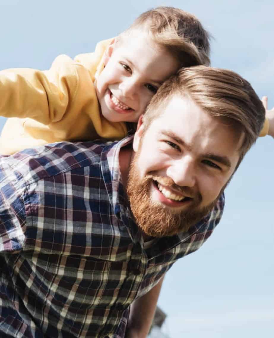 Ein Mann mit seinem Sohn auf dem Rücken lachen in die Kamera.